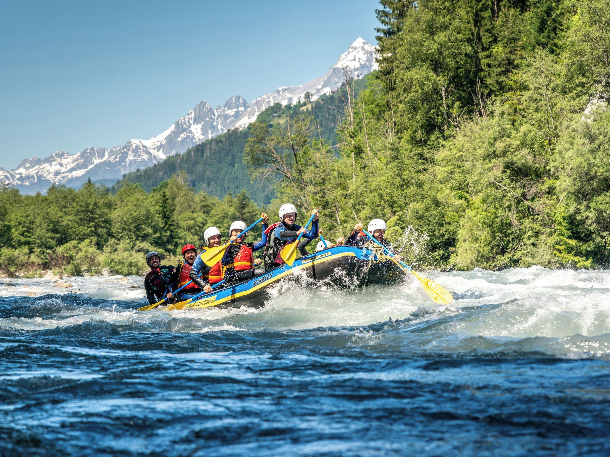 Un groupe de personnes à bord d’un radeau bleu et jaune navigue sur une rivière bordée de forêts et entourée de montagnes.