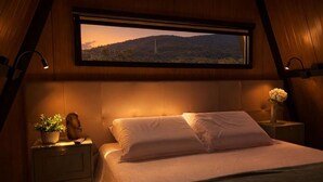 A bedroom with a bed, a nightstand with a plant, a vase with flowers, and a window with a view of a mountain. - Cabana Country Romântica com Banheira & Piscina (Palhoça)