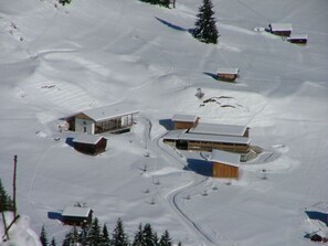 Une station de ski enneigée, composée de plusieurs bâtiments, dont un grand chalet et de plus petites cabanes, le tout entouré d’arbres et de pistes de ski.