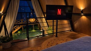 A cozy room with a bed, a TV displaying the Netflix logo, and a view of a wooden deck and garden at night. - Cabana Praiana - Vivendas Ferreira (Palhoça)