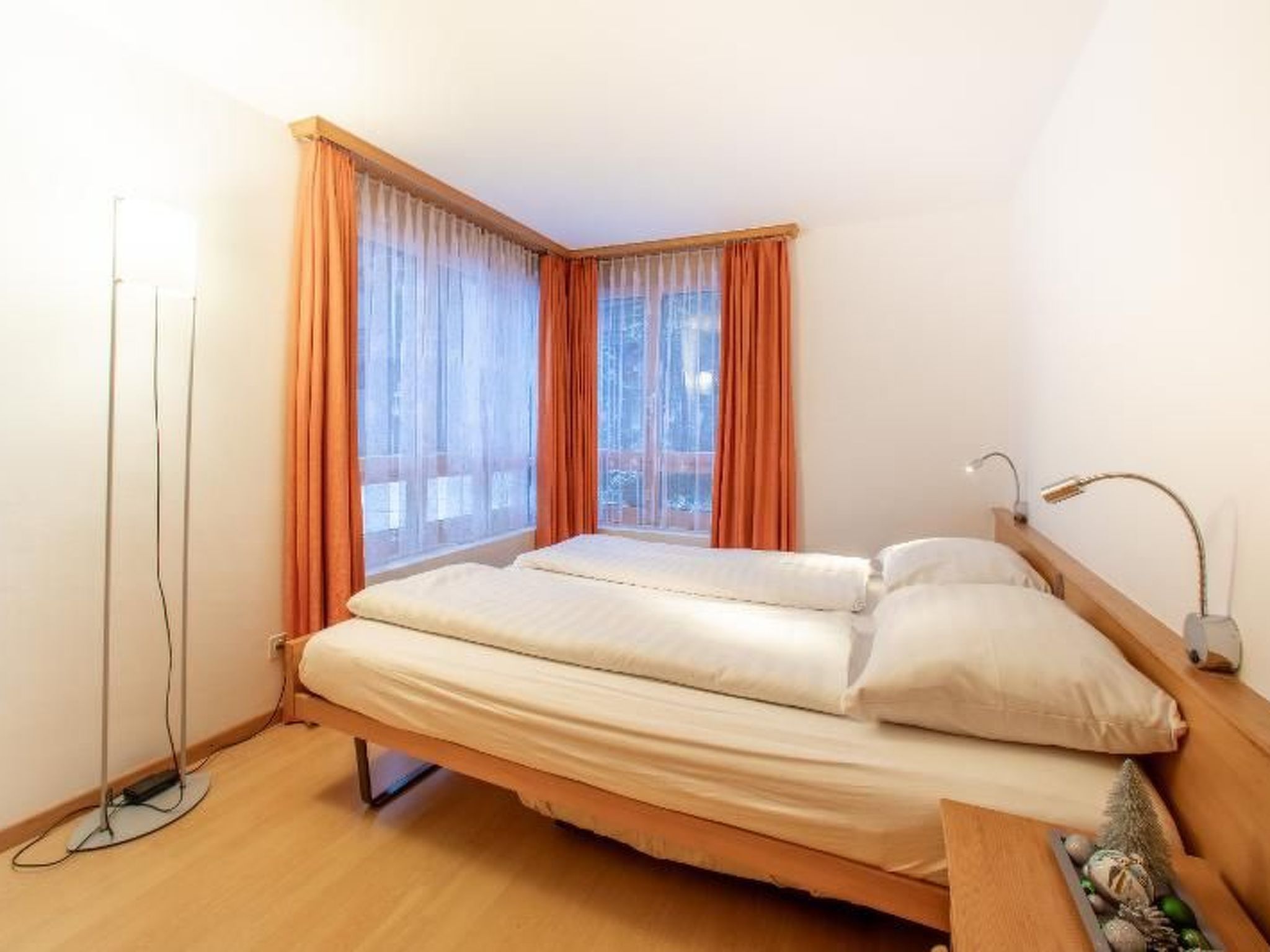 Un lit simple avec un cadre en bois et une literie blanche, dans une pièce dotée d’une grande fenêtre et de rideaux orange.