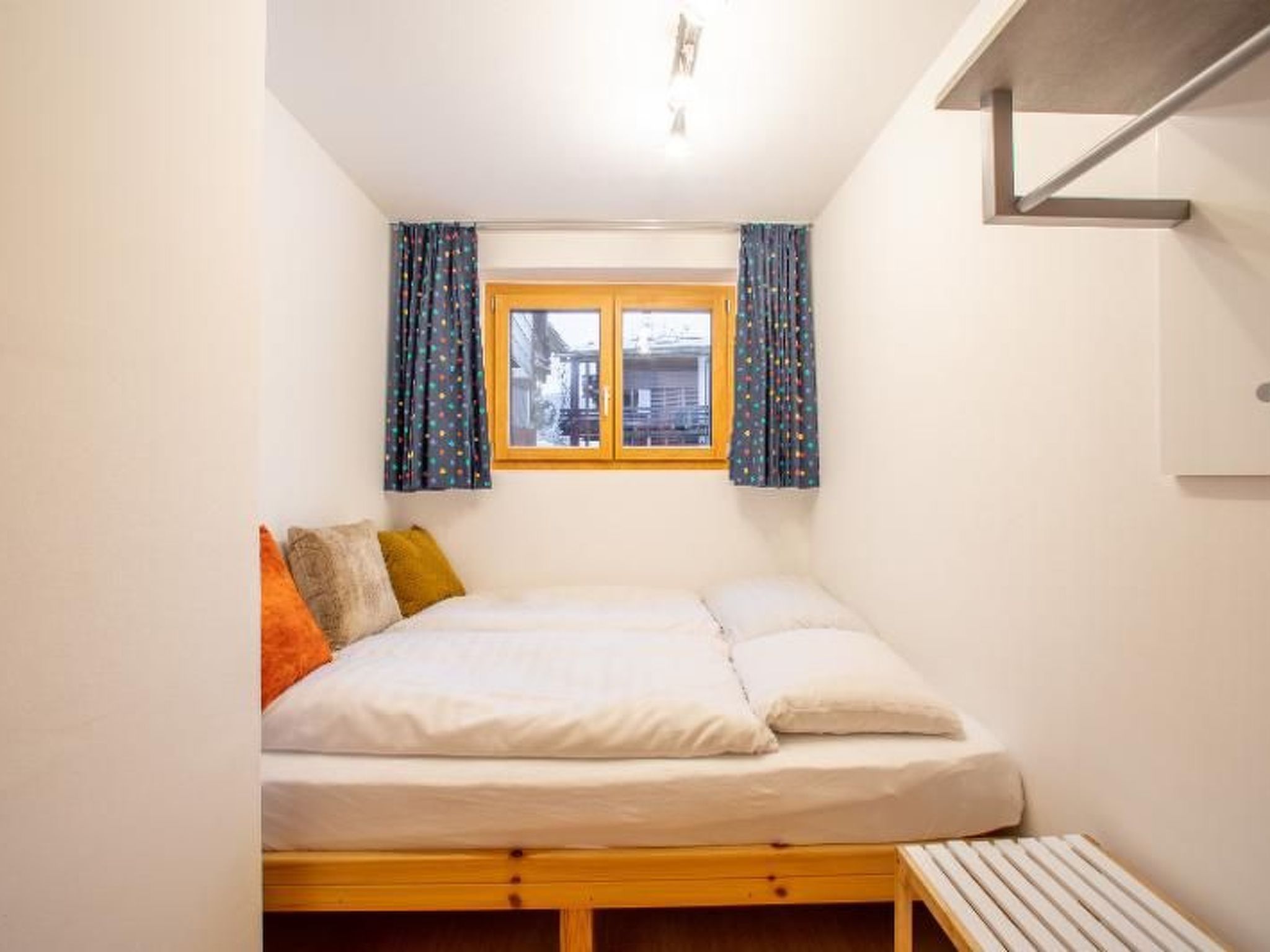 A small, single-bed room with a wooden bed frame, a window with curtains, and a bench at the foot of the bed.