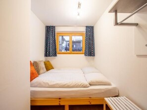 Ein kleines Zimmer mit einem Einzelbett, einem Holzbettgestell, einem Fenster mit Vorhängen und einer Sitzbank am Fußende des Bettes.