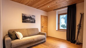 A room with a wooden ceiling, a beige sofa, a mountain landscape painting, a window with a view, and skis leaning against the wall. - Flem Mountain Lodge, (Flims Waldhaus), by Interhome (Flims)