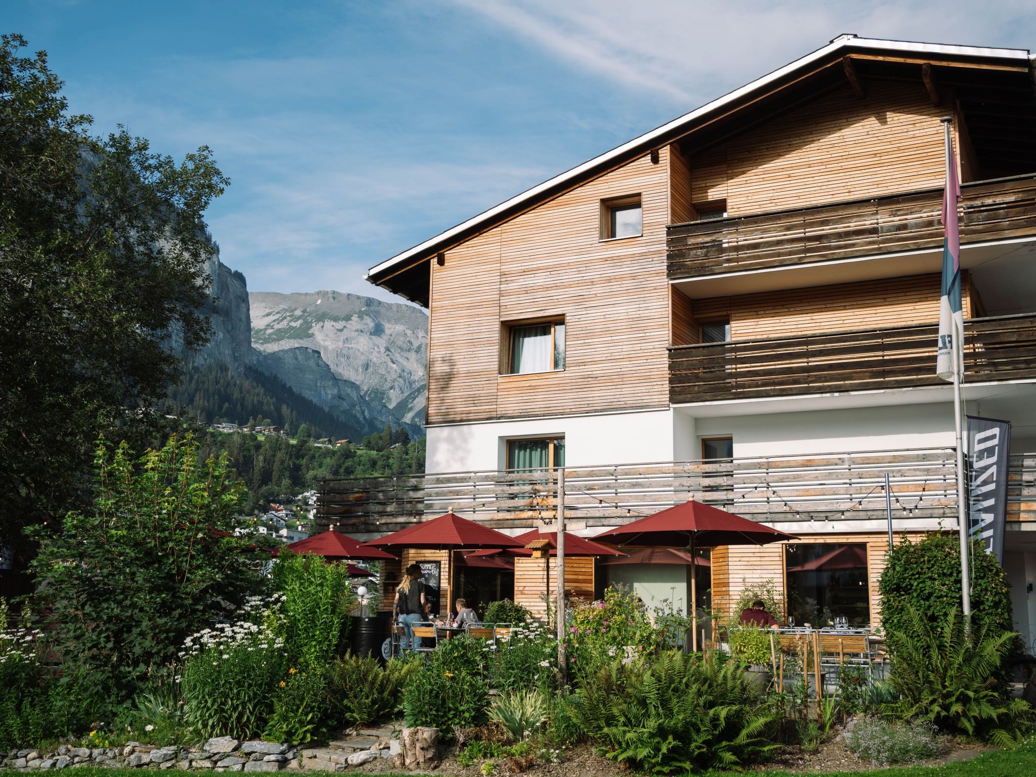 A two-story building with a wooden facade, outdoor seating with red umbrellas, and a mountainous backdrop.