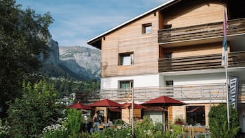 A two-story building with a wooden facade, outdoor seating with red umbrellas, and a mountainous backdrop.