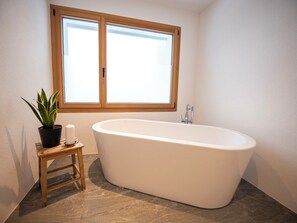 A bathroom with a freestanding bathtub, a window, a potted plant, and a small stool. - Flem Mountain Lodge, (Flims Waldhaus), by Interhome (Flims)
