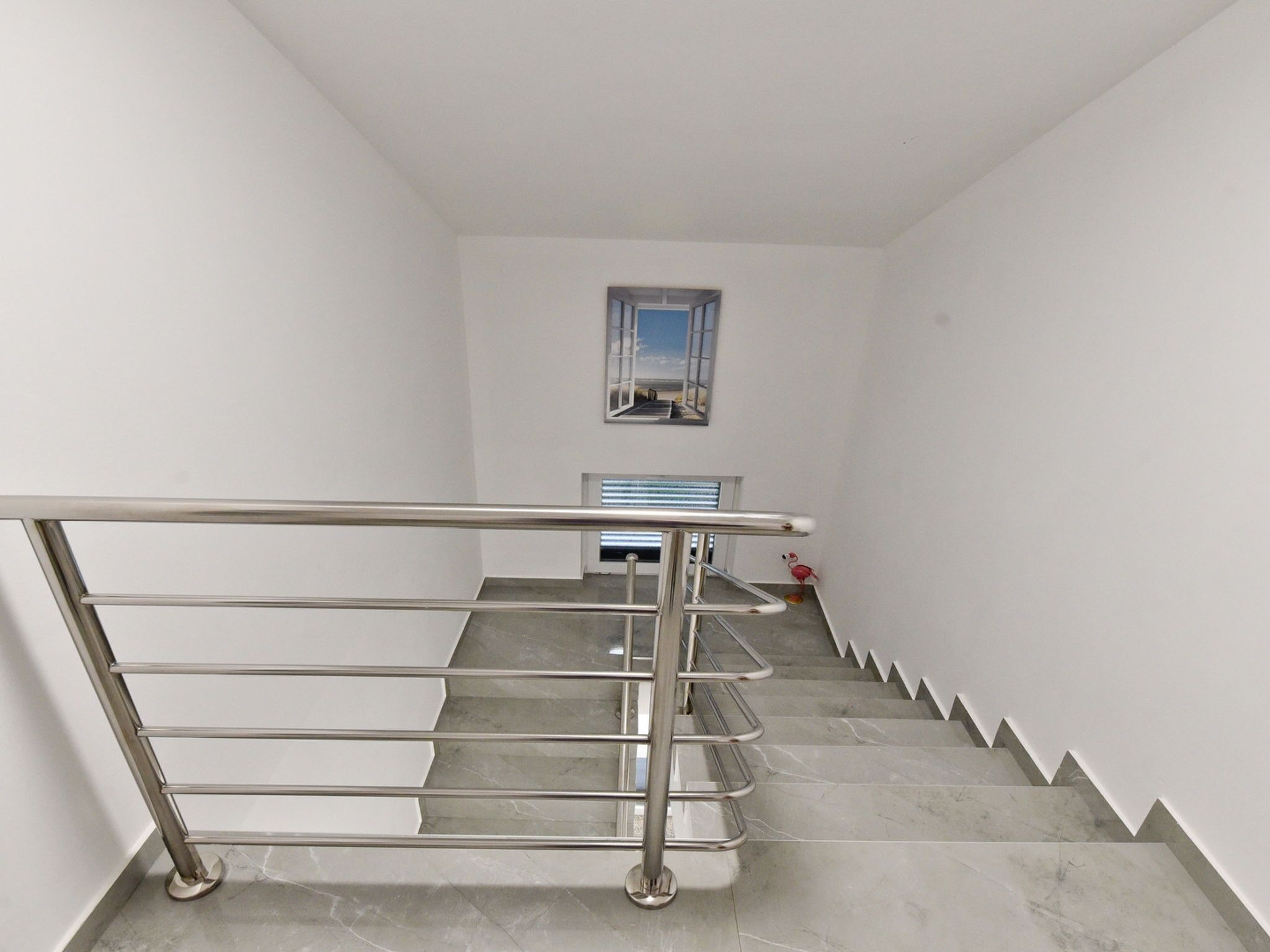 A staircase with a metal handrail, a small window with a view, and a red flamingo decoration.