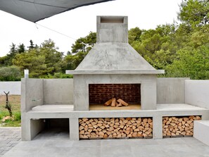 A concrete outdoor fireplace with a brick interior, surrounded by a low wall and a pile of firewood. - Vacation home  by Interhome (Crno)