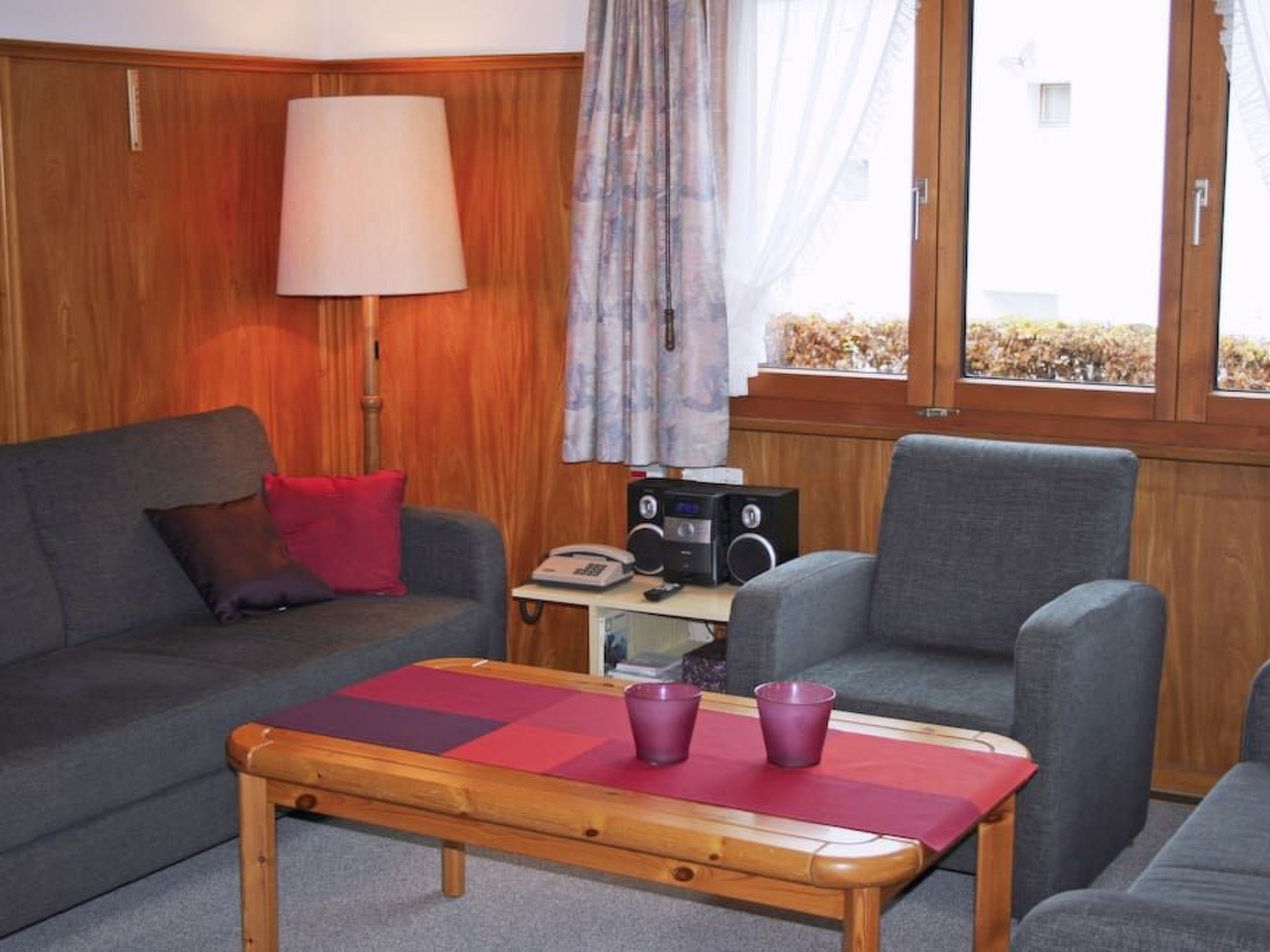 A room with a wooden paneled wall, a grey sofa, a wooden coffee table with a red placemat, a grey armchair, a lamp, and a window with curtains.
