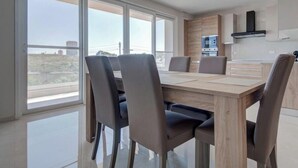 A modern dining area with a large table and chairs, large windows, and a kitchen in the background. - Pembroke 2BR Sea View Walk to Beach & Town (Pembroke)