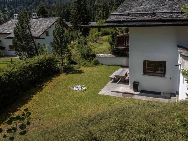 Un bâtiment blanc avec un toit en pierre, un petit espace extérieur aménagé avec des sièges et une cour verdoyante.