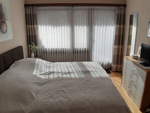 Une chambre à coucher avec un lit, une commode, un téléviseur et une fenêtre avec des rideaux.