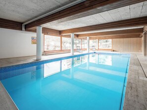 Ein Hallenbad mit klarem, blauem Wasser, Holzbalken an der Decke und großen Fenstern mit Blick auf Bäume.