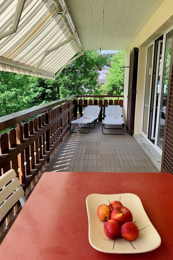 A covered outdoor seating area with tables and chairs, a view of greenery, and a table with a plate of fruit.