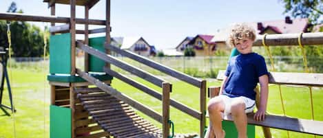 Un niño sentado en una estructura de juego de madera con un tobogán, rodeado de césped verde y casas al fondo.