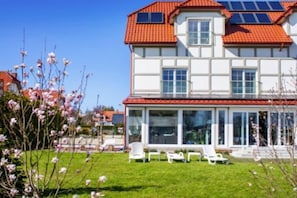 Een twee verdiepingen tellend huis met een rood tegeldak, grote ramen en een overdekt terras met buitenmeubilair.