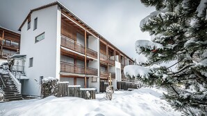 Un immeuble à plusieurs étages doté d’une façade en bois, d’un espace extérieur couvert et entouré d’un paysage enneigé.