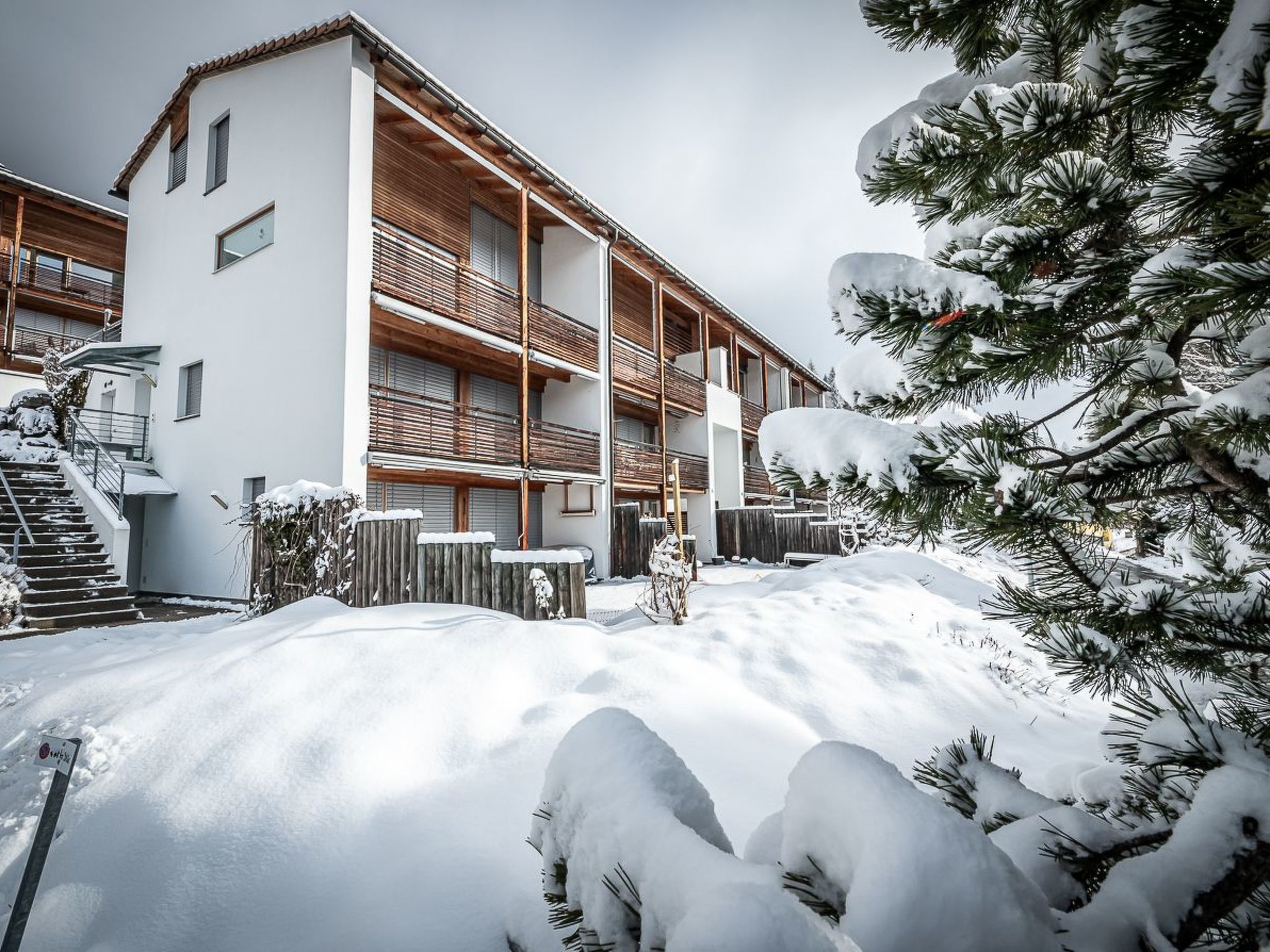A multi-story building with a wooden balcony, surrounded by snow-covered ground and trees.