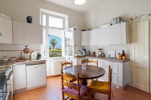 A kitchen with white cabinets, a dining table with chairs, a window with a view, and a radiator. - A charming, detached, 200-square-meter period house on three levels, accommodating up to 10 people.The property is located in Gravedona e Uniti, a charming village on the northwest shore of Lake Como, known for its historic center and picturesque lakefro (Gravedona ed Uniti)