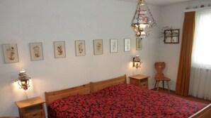 A bedroom with a wooden bed, a nightstand, a chair, a wall-mounted lamp, and framed pictures on the wall. - Ner B / Hartl, (Flims Dorf), 29 by Interhome (Flims)