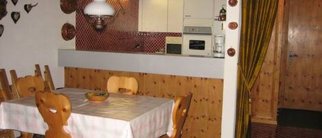 Uma área de refeições com mesa posta para quatro pessoas, cadeiras de madeira, tapete estampado e uma cozinha acima, com armários e micro-ondas.