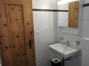 A bathroom with a wooden door, white tiled walls, a white sink with a mirror above it, and a small trash bin. - Ner B / Hartl, (Flims Dorf), 29 by Interhome (Flims)