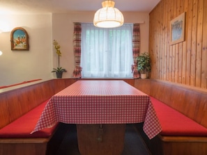 A dining area with a table covered in a checkered tablecloth, wooden booth seating, a hanging light fixture, and a window with curtains. - Casa Mirella, (Flims Dorf), 456 by Interhome (Flims)