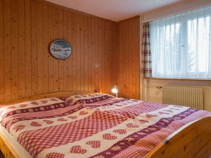 A wooden-paneled room with a bed, a round wall decoration, and a window with curtains. - Casa Mirella, (Flims Dorf), 456 by Interhome (Flims)