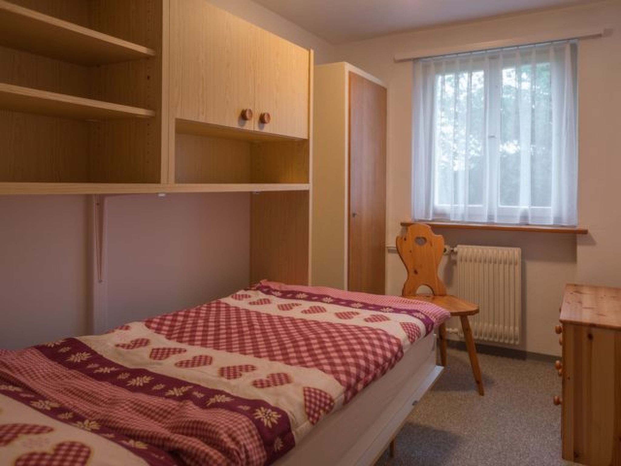 A bedroom with a bed, a wooden chair, a window with curtains, and built-in shelves.