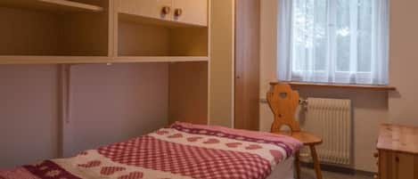 Una camera da letto con un letto, una sedia in legno, una finestra con tende e scaffali a muro.