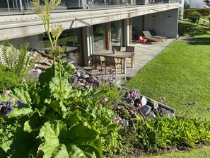 A modern outdoor patio with a dining table and chairs, a seating area with sun loungers, and a garden with various plants and flowers. - Casa Admisa, (Flims Dorf), 2623 by Interhome (Flims)
