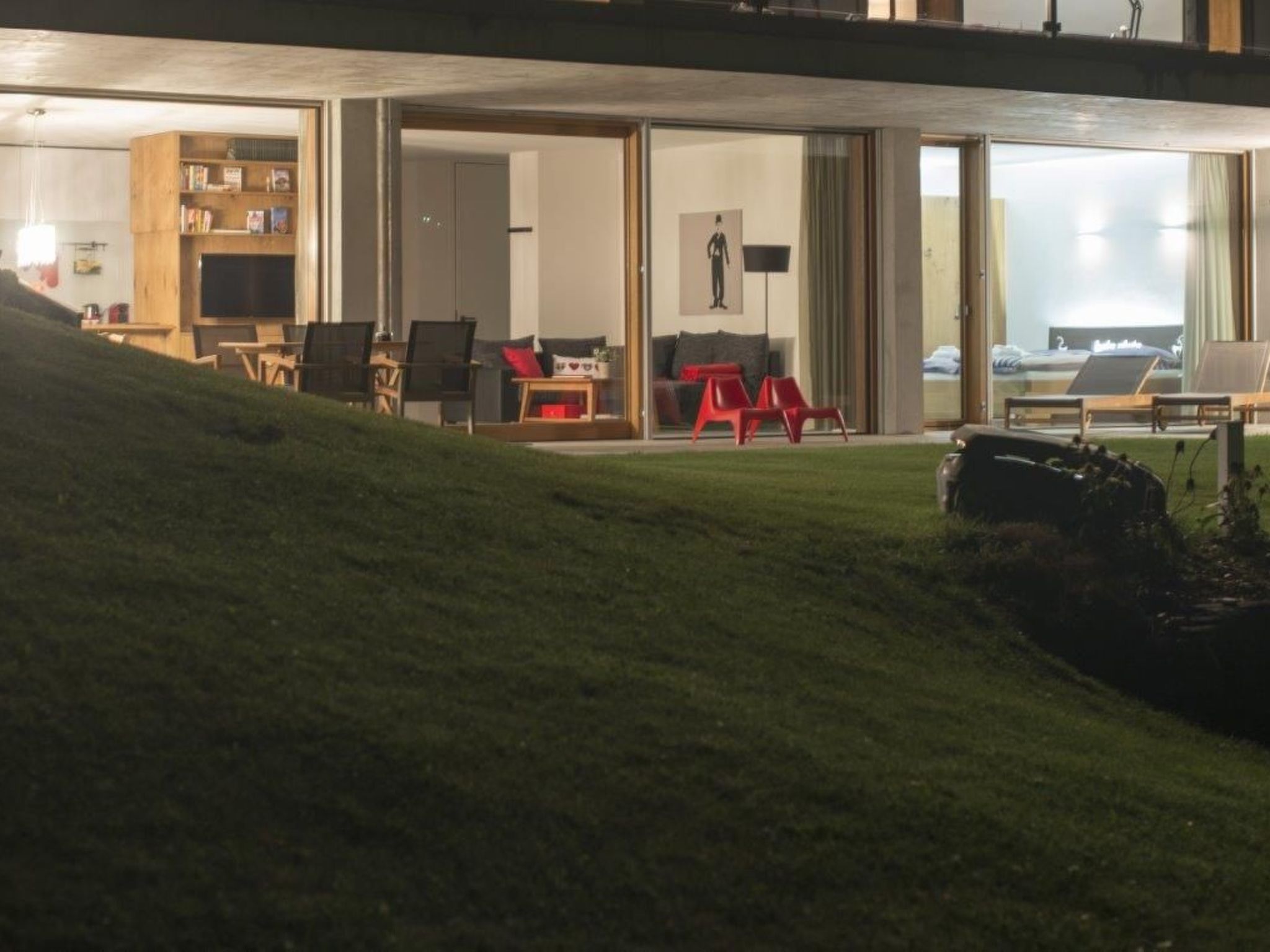 Ein modernes Hotelzimmer mit einer großen Schiebeglasdoor zu einem Balkon, einer Sitzecke mit Stühlen und einem roten Hocker sowie einem Bücherregal mit Büchern und Dekorationsobjekten.