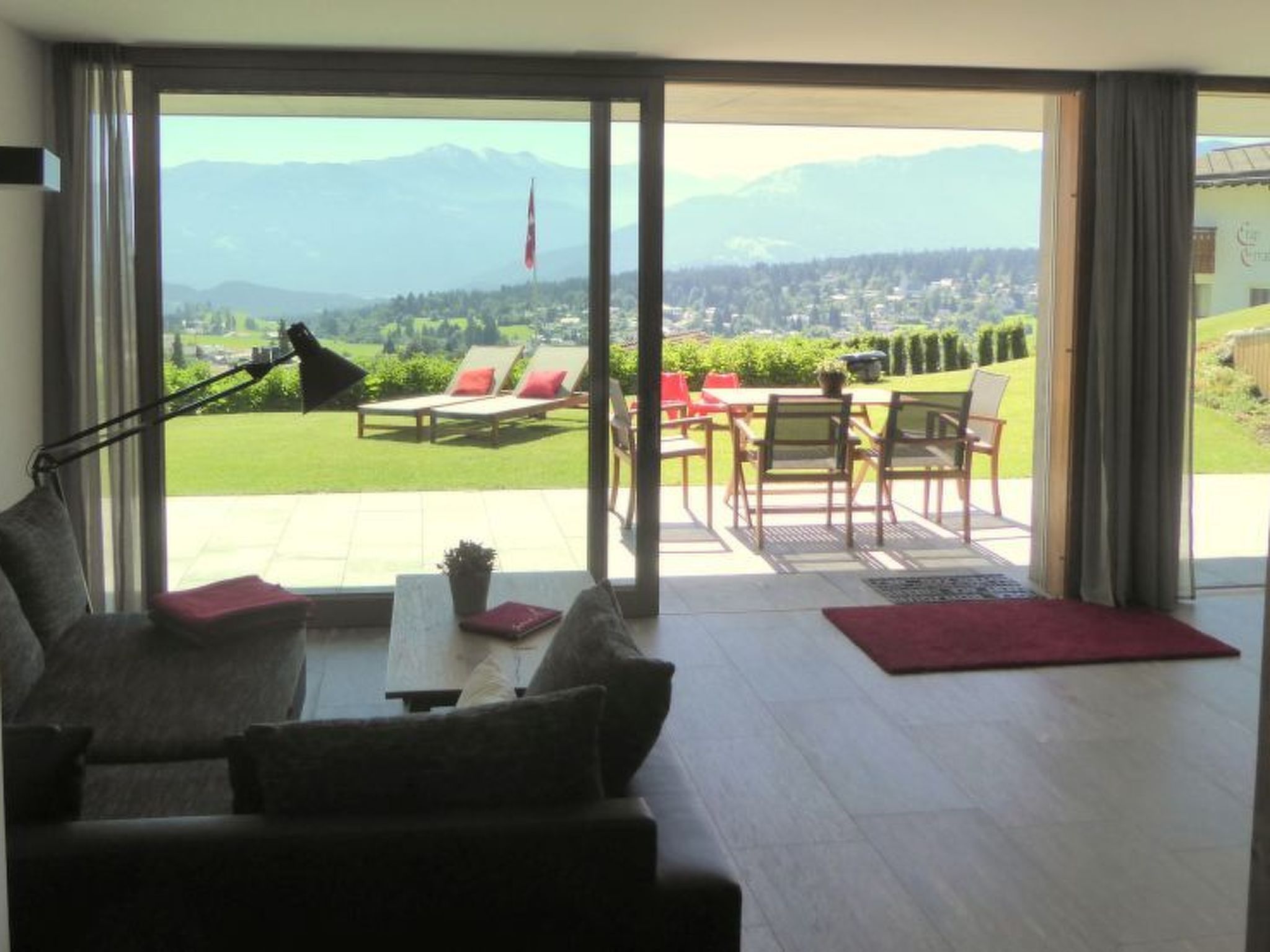 A room with a sofa, a table, and a view of a garden and mountains through large glass doors.