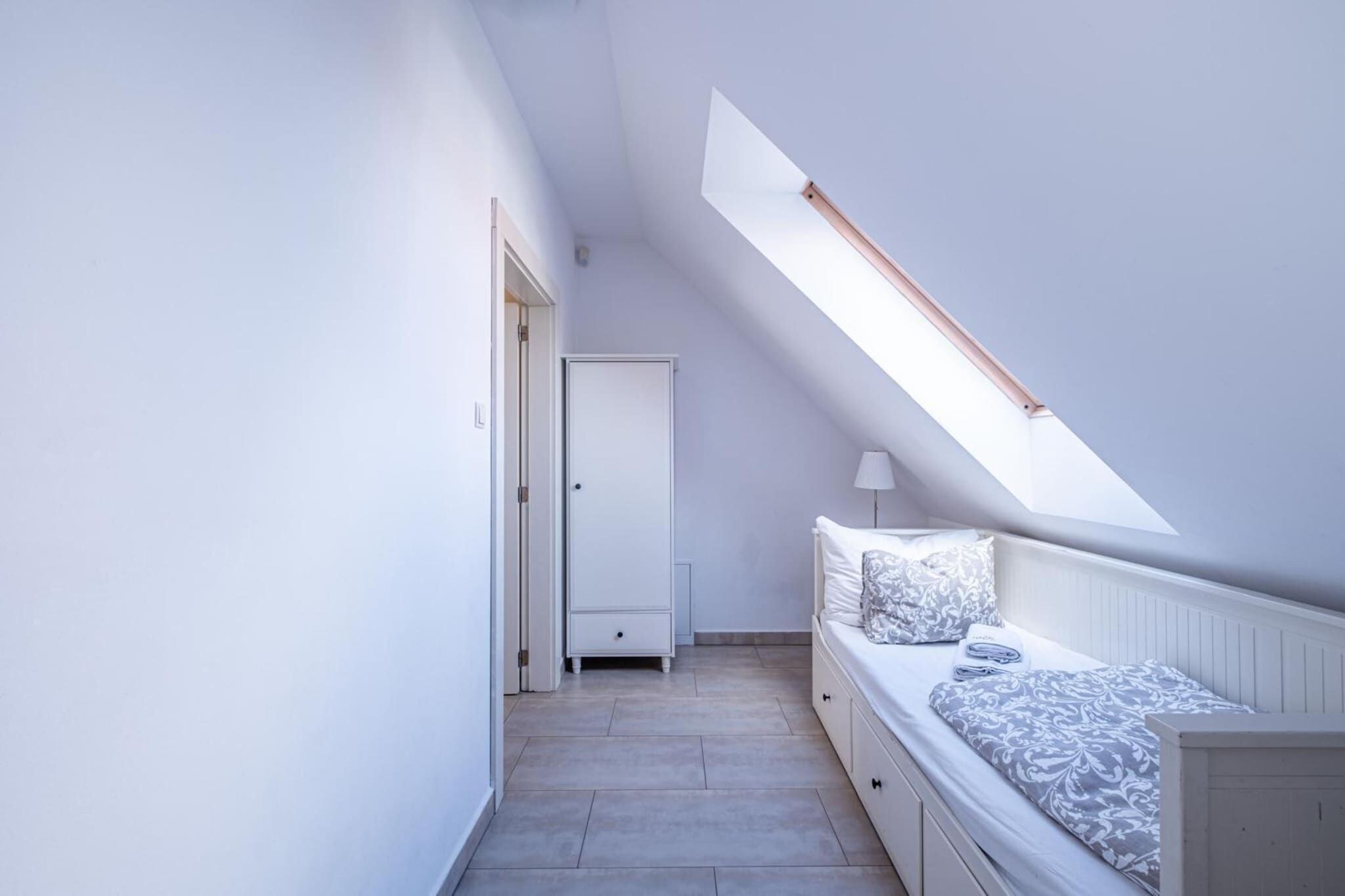 Ein schmales, gut beleuchtetes Zimmer mit schrägem Dach, einem Bett mit gemusterten Textilien, einem Nachttisch und einer Tür zu einem weiteren Raum.
