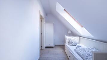 Ein schmales, gut beleuchtetes Zimmer mit schrägem Dach, einem Bett mit gemusterten Textilien, einem Nachttisch und einer Tür zu einem weiteren Raum.