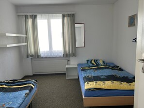 Une chambre avec deux lits, une fenêtre avec des rideaux, un radiateur, une petite étagère et un miroir fixé au mur.