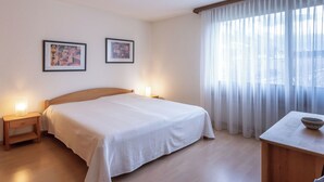 Una habitación de hotel con una cama, mesitas de noche, dos cuadros enmarcados, un ventanal con cortinas translúcidas y una pequeña mesa con un cuenco.