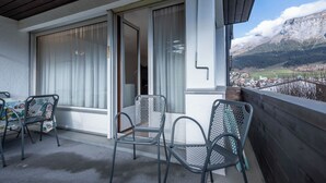 Un balcón con vistas a un valle y montañas, una mesa con sillas y un ventanal corredera.