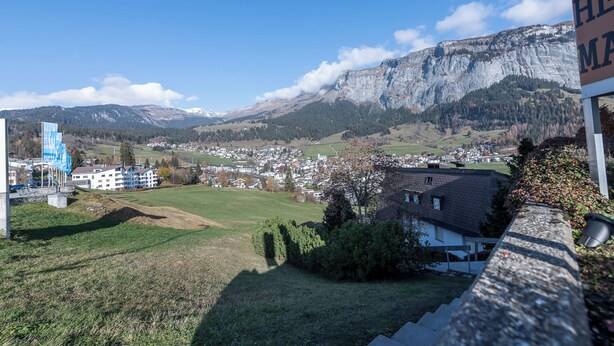 Un pueblo con una montaña de fondo, una zona verde y un edificio con un cartel.