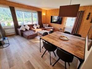 Ein modernes Wohnzimmer mit Holzboden, einer Sofagarnitur, einem Esstisch mit Stühlen, einem an der Wand montierten Fernseher und großen Fenstern mit Vorhängen.