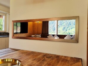 Ein modernes Interieur mit einem großen Spiegel, der einen Raum widerspiegelt, einem Holztisch und einem Fenster mit Blick auf Bäume.