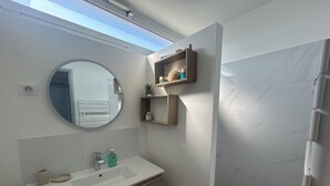A bathroom with a round mirror, a sink, wooden cabinets, and a shelf with toiletries. - Sea view apartment, quiet residence, 10 minutes from the center and beaches. (Bandol)