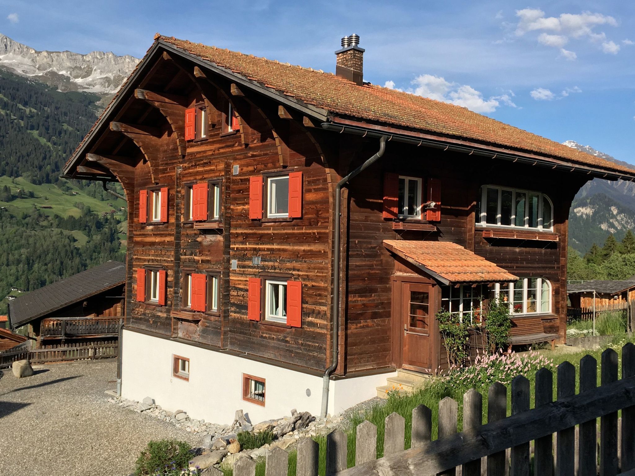 A multi-story wooden chalet with red shutters, set against a mountainous backdrop.