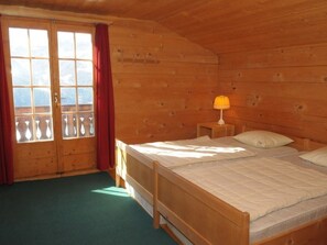 Ein Zimmer mit Holzboden, einem Bett, einem Nachttisch, einem Fenster mit Vorhängen und Blick auf die Berge.