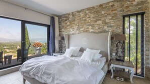 A bedroom with a large bed, stone wall, a view of the landscape, and a small table with a lamp. - Villa with Pool and Spectacular Sea Views (Sa Riera)