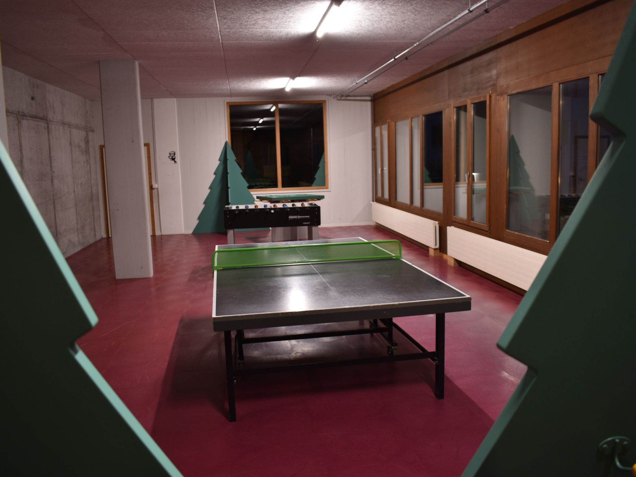 Een tafeltennistafel in een ruimte met een rode vloer, een raam en een houten kast.