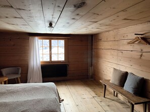 Une pièce aux murs en bois, une fenêtre avec des rideaux, un radiateur, un lit, un banc et une chaise.