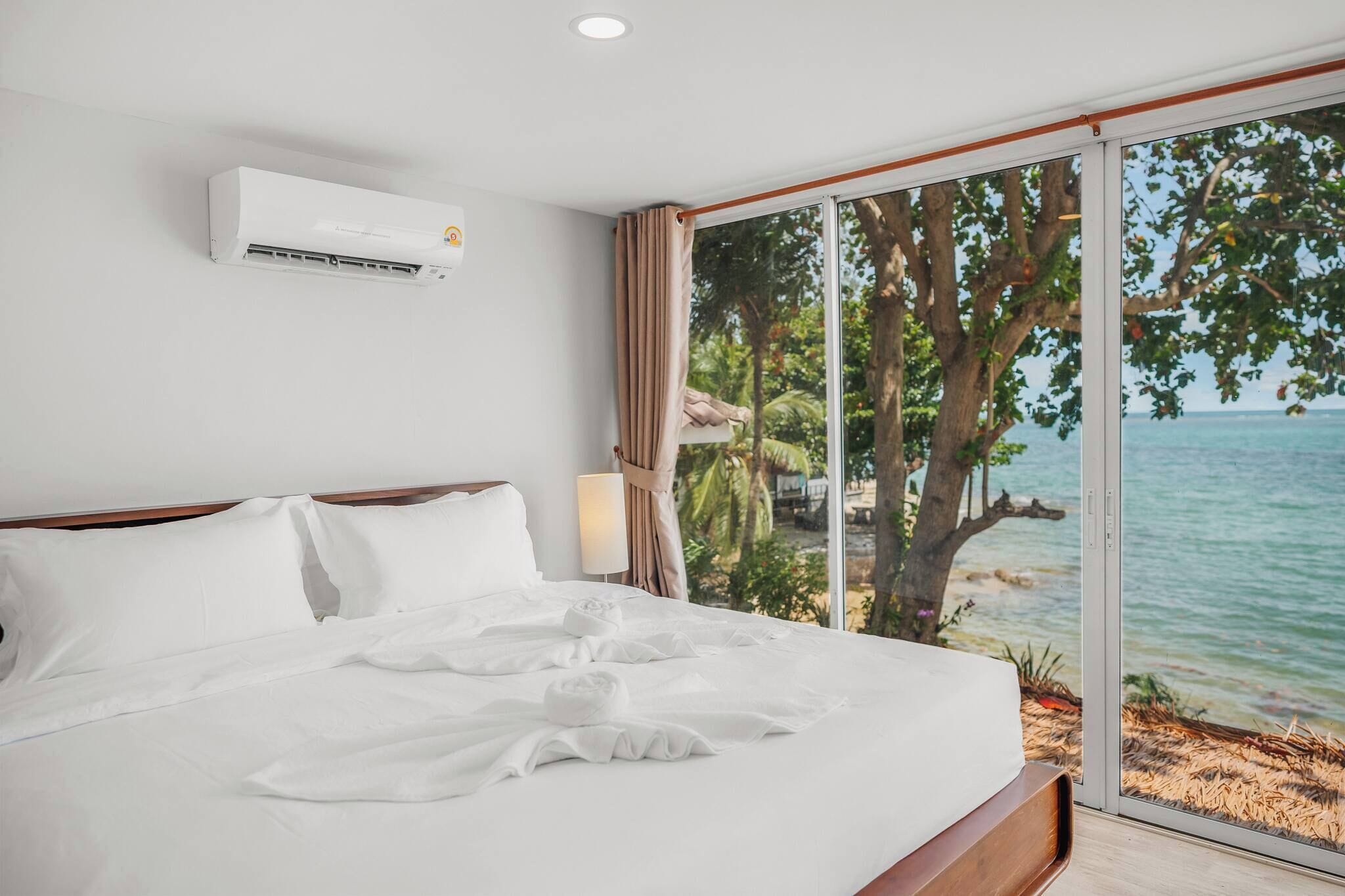 A hotel room with a large bed, an air conditioning unit, a view of the sea, and a window with curtains.