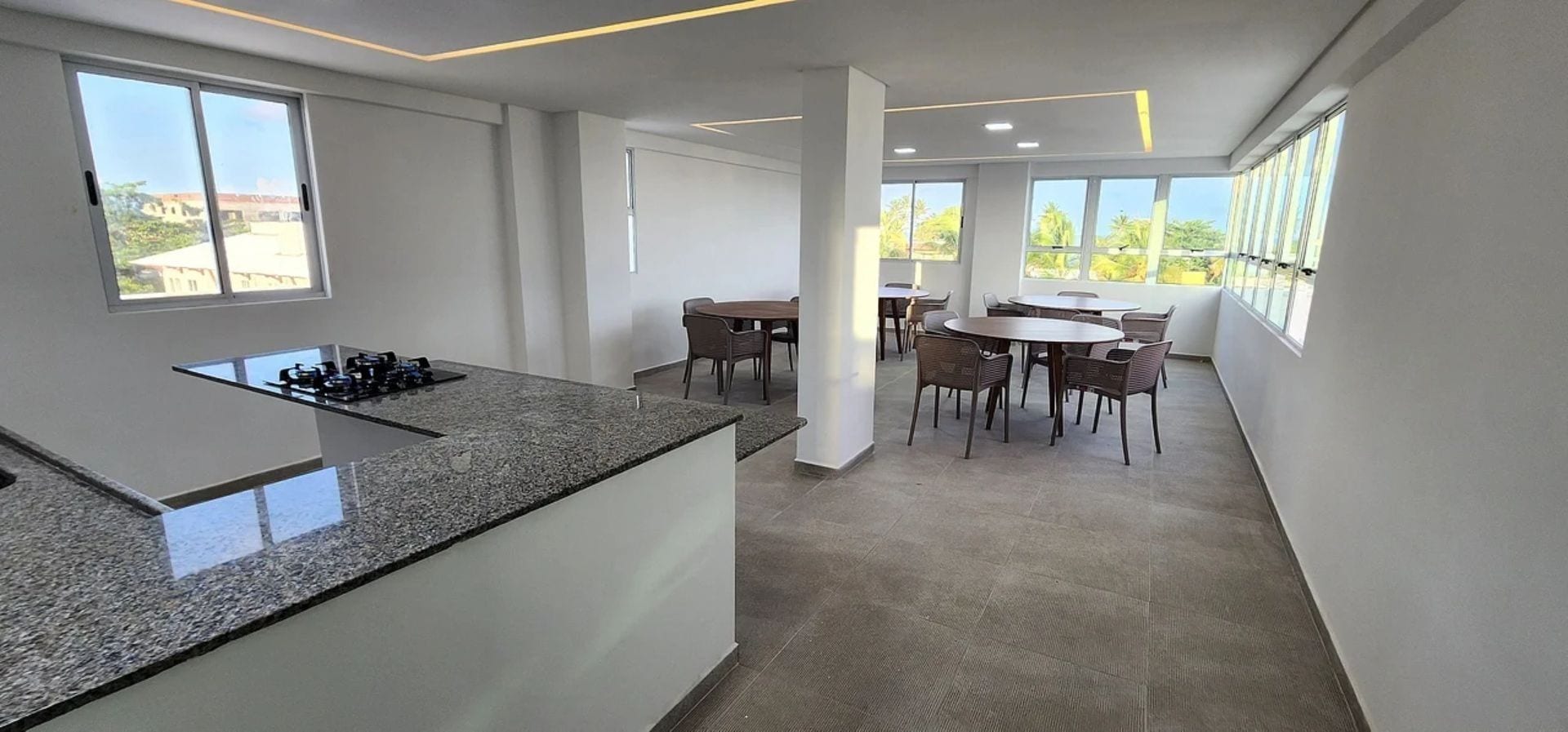 A modern kitchen with a granite countertop and a built-in stove. There are multiple dining tables with chairs arranged in the dining area. Large windows provide natural light and views of the outside.